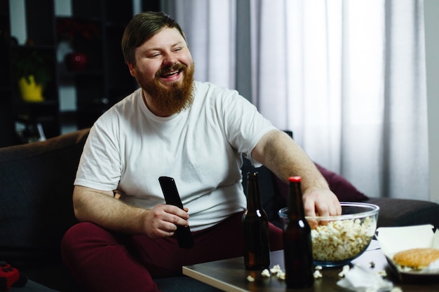 Happy fat man sits on the sofa and watches TV with popcorn and beer Free Photo