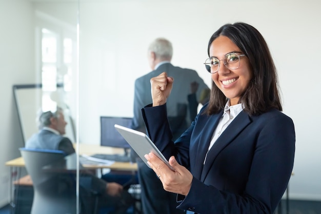Happy female professional in glasses and suit holding tablet and making winner gesture while two businessmen working behind glass wall. copy space. communication concept Free Photo