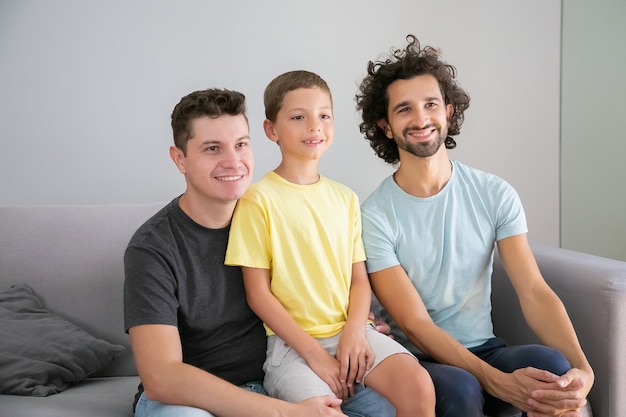 幸せなゲイの父親と子供が自宅のソファに座って 笑顔で 目をそらしています 正面図 家族と親の概念 無料の写真