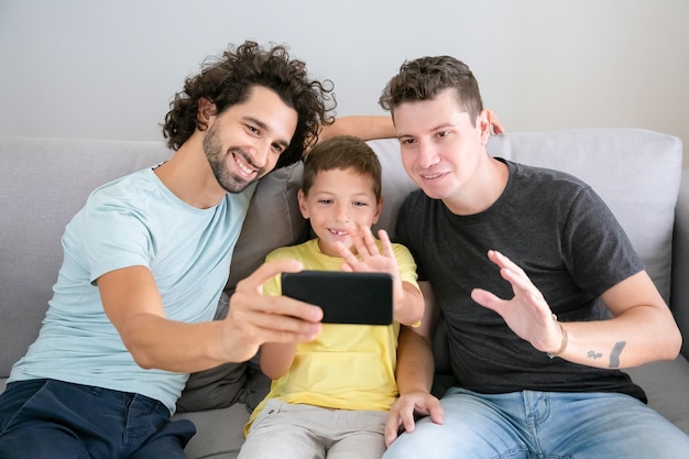 幸せなゲイの両親と子供がビデオ通話に携帯電話を使用し 自宅のソファに座って 正面カメラで手を振って 笑っています 正面図 家族とコミュニケーションの概念 無料の写真