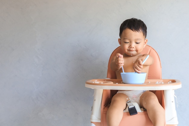 Happy infant asian baby boy sitting on baby high chair and eating food by himself with copy space. P
