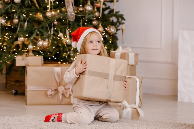自宅のクリスマスツリーの近くにクリスマスギフトボックスとサンタの帽子で幸せな小さな笑顔の女の子 プレミアム写真