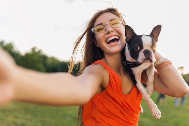 自撮り写真を作る幸せなきれいな女性公園 ボストンテリア犬を抱いて ポジティブな気分を笑顔 トレンディな夏のスタイル オレンジ色のドレス サングラスを身に着けて ペットと遊んで 楽しんでください 無料の写真