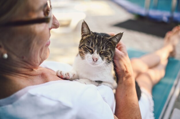 国内のぶち猫を抱き締める屋外の夏の庭でリラックスしたメガネで幸せな笑顔の年配の女性 引退した老人と動物のペットの概念 プレミアム写真