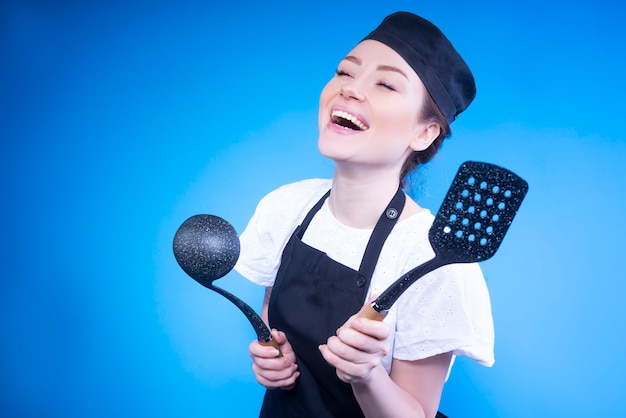 Happy woman chef laughing and holding kitchen utensils in her hands against blue wall Free Photo