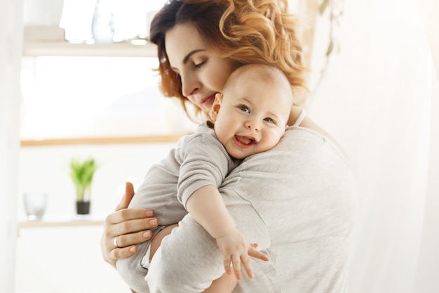 Happy young mom holds precious little child and gently hugging his little body. kid laughing joyfully and looking at camera with big grey eyes. Free Photo