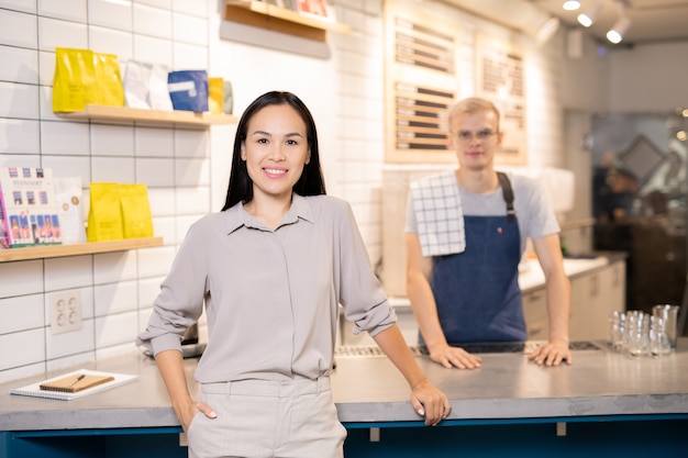 背景に制服を着たウェイターとカメラの前のカウンターのそばに立っているレストランやカフェの幸せな若い所有者 プレミアム写真