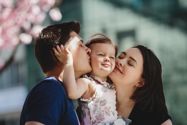 Happy young parents with a little daughter stand under blooming pink tree outside Free Photo