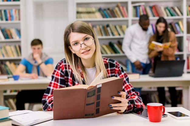 Happy young woman in casual wear and eyeglasses sitting in the library with book, searching information for her studies. Premium Photo