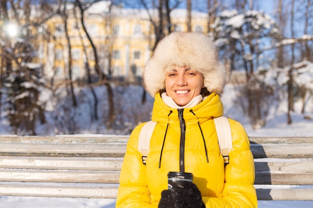 晴れた日に雪の公園で暖かい服を着て冬の幸せな若い女性はベンチに座って 新鮮な空気とコーヒーを一人で楽しんでいます 無料の写真