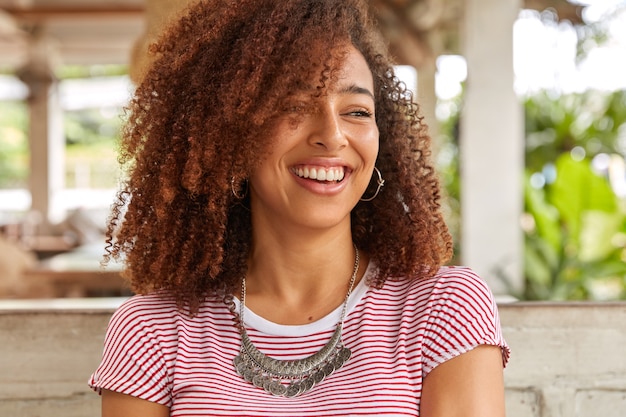Free Photo Headshot Of Funny Black Woman With Curly Hair Laughs At Good Joke Has Toothy Smile Shows White Perfect Teeth Wears Striped Casual T Shirt Poses At Terrace Cafeteria