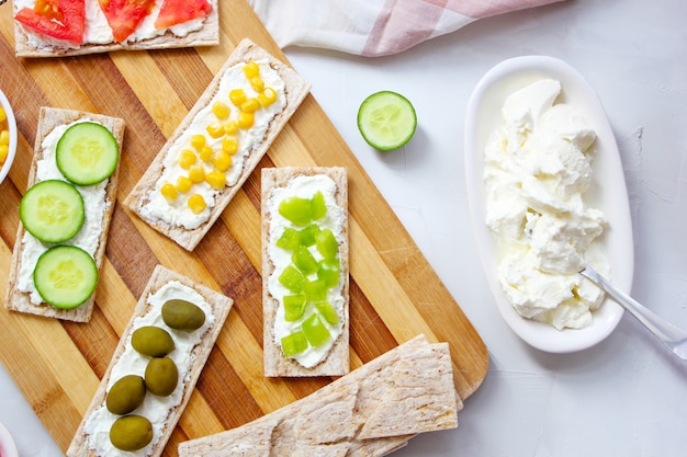 Homemade crispbread toast with cottage cheese and green olives, slices of cabbage, tomatoes, corn, green pepper on cutting board. healthy food concept, top view. flat lay Free Photo