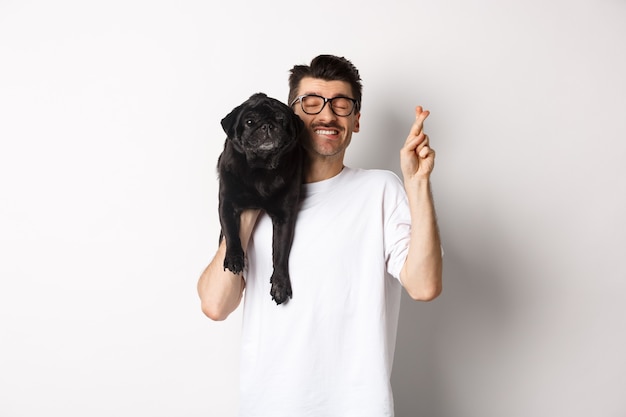 Premium Photo Hopeful Smiling Dog Owner Making A Wish Holding Cute Black Pug On Shoulder And Cross Fingers For Good Luck White Background