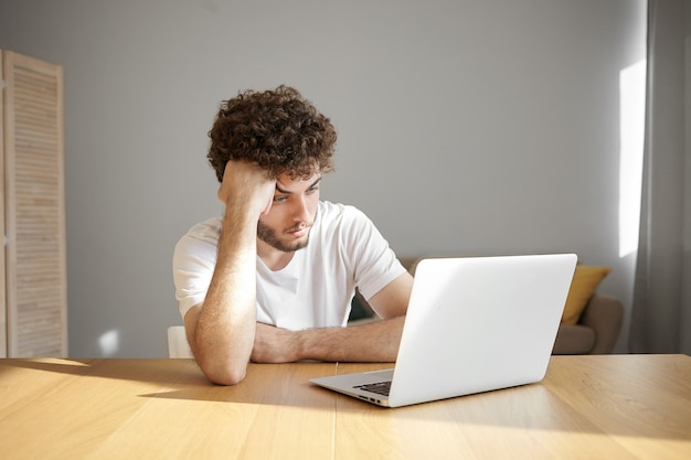 Horizontal shot of frustrated young unshaven male freelancer in white t-shirt using electronic device for distant work, having tired or bored look while working on urgent project early in the morning Free Photo