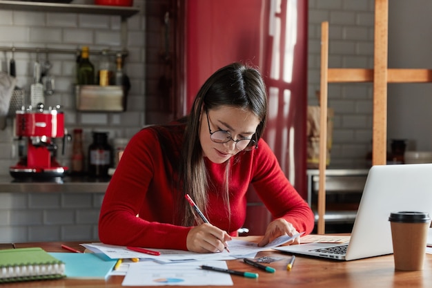 Horizontal shot of pleasant looking businesswoman works with papers at home, prepares report document, fills in information, sits in front of opened laptop at kitchen. Free Photo