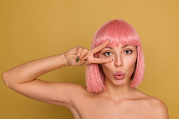 Horizontal shot of pretty young blue-eyed lady with short pink hair holding victory gesture near her face and folding her lips together while posing over mustard wall Free Photo