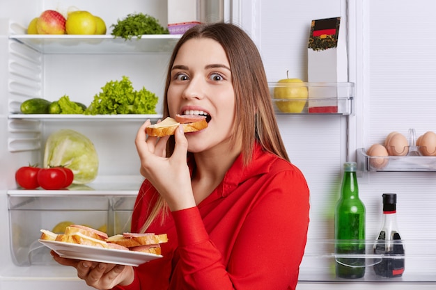 Premium Photo | Hungry adorable woman eats delicious sandwhiches with  cheese and sausage, stands near opened refrigerator full of products.  beautiful female feels hunger after work. people and eating concept