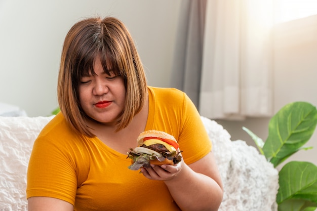 ハンバーガーを笑顔で押しながら寝室に座っている空腹の太りすぎの女性 彼女はとても幸せで ファーストフードを食べるのを楽しんでいます 過食症 Bed の概念 プレミアム写真