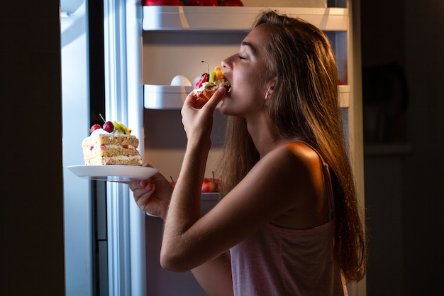 冷蔵庫の近くで夜に甘いケーキを食べるパジャマ姿の空腹の女性。高炭水化物食品と不健康な夜の食事のために、食事を止めて余分な体重を増やします Premium写真