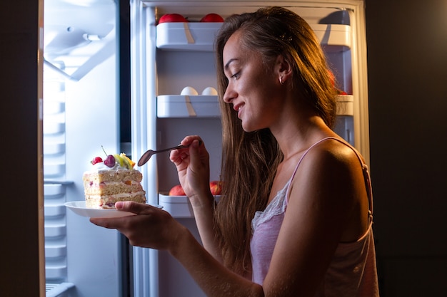 Hungry woman in pajamas eating sweet cake at night near fridge. stop diet and gain extra pounds due to high carbs junk food and unhealthy night eating Premium Photo