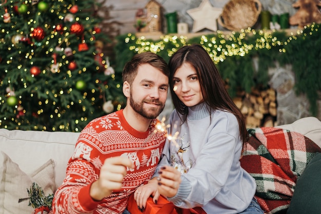 家でクリスマスを祝っている間 線香花火を持って 抱き締めて 笑顔でセーターを着て幸せな若いカップルの画像 心地よさとお祭りの雰囲気 プレミアム写真