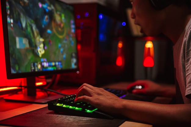 暗い部屋でコンピューターでビデオゲームをプレイし ヘッドフォンを着用し バックライト付きのカラフルなキーボードを使用して興奮しているアジアの10代の ゲーマー少年の画像 プレミアム写真
