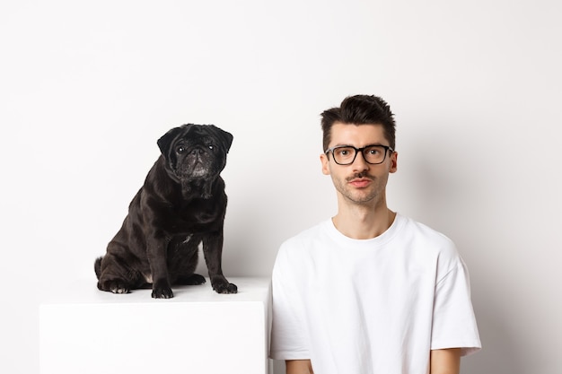 黒のかわいいパグ犬の隣に座っている眼鏡をかけたハンサムなヒップスターの画像 どちらも白い背景の上でカメラを見つめています 無料の写真