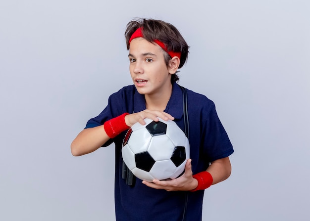 ヘッドバンドとリストバンドを身に着けている印象的な若いハンサムなスポーティな少年は コピースペースで白い背景にまっすぐに孤立して見えるサッカーボールを保持している首の周りに歯列矯正器と縄跳びを身に着けています 無料の写真