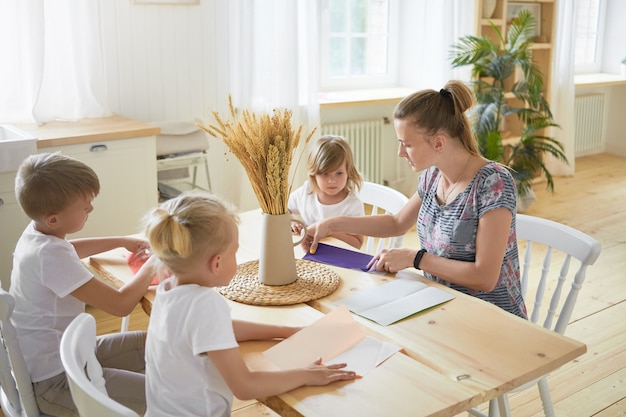 広々としたリビングルームのダイニングテーブルに座って 子供 たちに折り紙の作り方を教える若い女性のベビーシッターの屋内画像 家で母親と一緒に紙飛行機を作る3人の子供 無料の写真