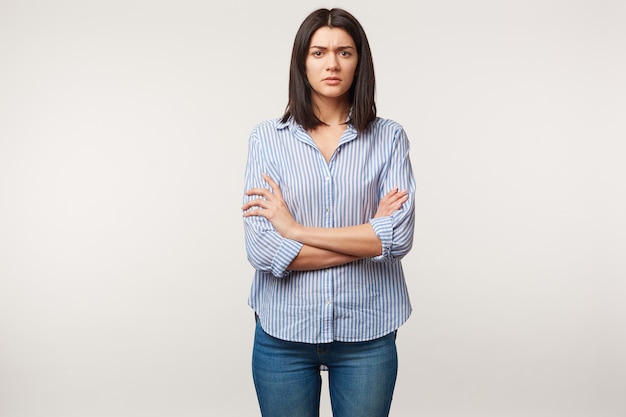 Free Photo Indoor Photo Of Brunette Woman Looks With Distrust Wary Suspicion Tense Listens To Someone With Doubt Stressful Standing With Arms Folded Dressed In Jeans And Striped Shirt Over White