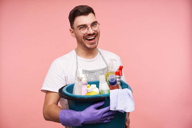 Indoor portrait of glad young unshaved brunette man in uniform preparing for spring cleaning and holding household chemicals while posing on pink Free Photo