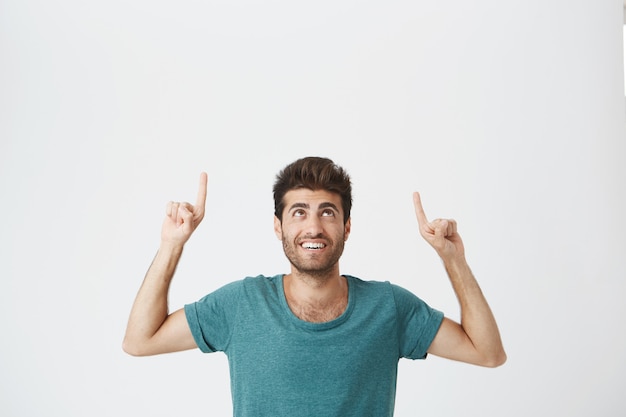 Free Photo Indoor Portrait Of Joyful Bearded Spanish Guy With Pleased Expression Wearing Blue Tshirt Laughing And Pointing Upside On White Wall Copy Space