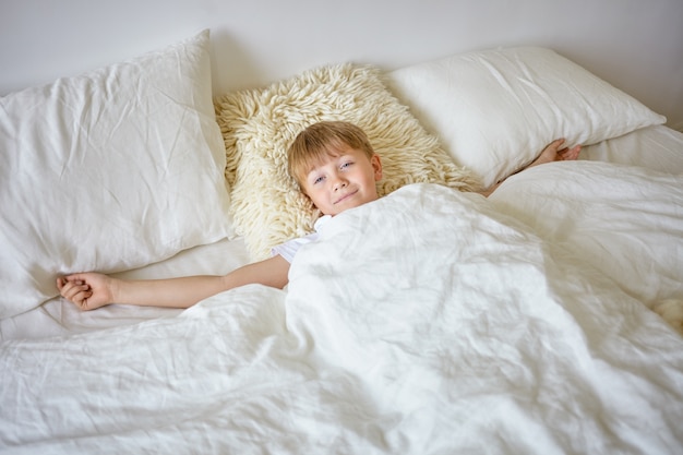 Indoor portrait of sleepy european teenage boy stretching arms after awakening early in the morning, lying on white bedclothes, going to school, looking , having lazy facial expression Free Photo