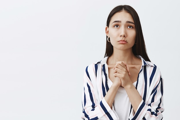 Indoor shot of gloomy cute female in striped blouse over t-shirt, clasping hands and pursing lips, frowning while gazing up with hopeful expression, waiting for results of test over gray wall Free Photo