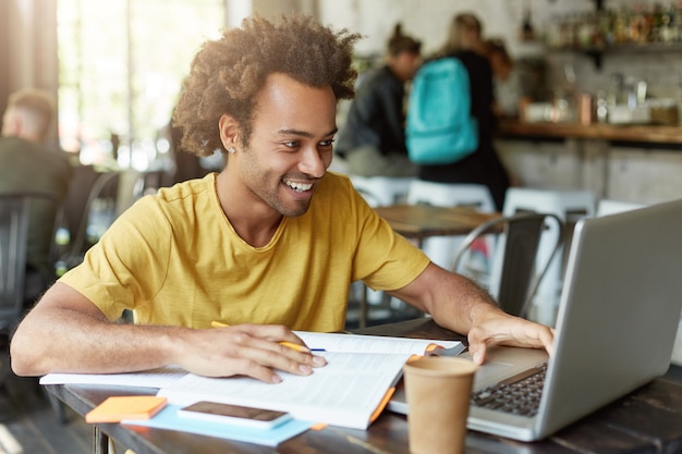 Indoor shot of happy student male with curly hair dressed casually sitting in cafeteria working with modern technologies while studying looking with smile in notebook receiving message from friend Free Photo