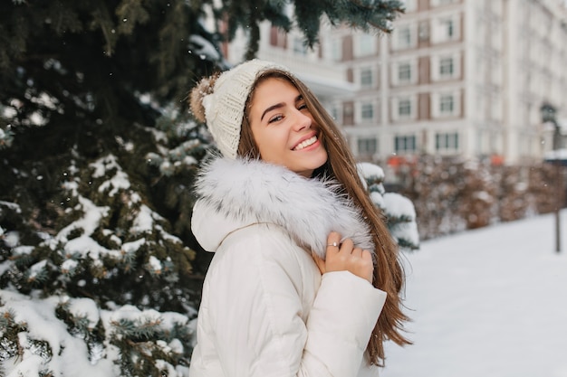 幸せな冬の時間を楽しんで笑っている白い服装で興味のある長髪の女性 雪に覆われた通りにニット帽子立っている壮大なヨーロッパの女性の屋外のポートレート 無料の写真