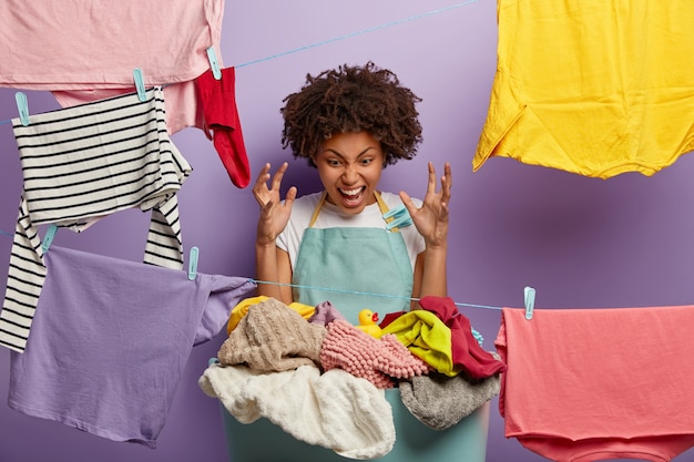 Irritated dark skinned woman raises arms, looks angirly at pile of dirty linen, doesnt want to wash clothes by hands as washing machine is broken, hates laundry process, wears apron with clothespins Free Photo