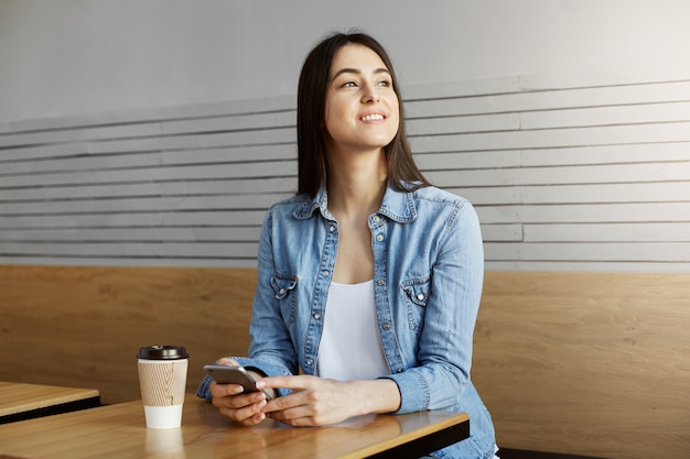 カフェに座っている黒髪のうれしそうな魅力的な女の子は コーヒーを飲み スマートフォンで友達とチャットしてから 頭を回して窓からボーイフレンドを見る 無料の写真