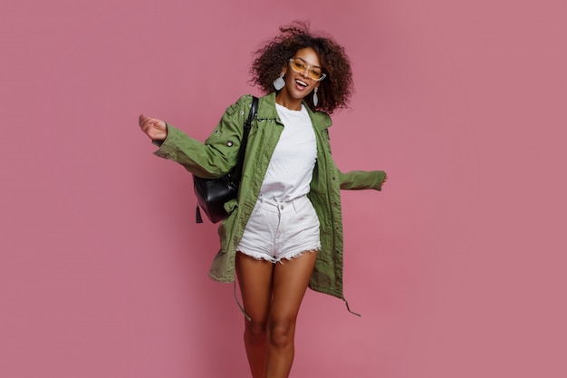 Joyful black woman having fun in studio over pink background. white t-shirt, green jacket. stylish spring look. Free Photo