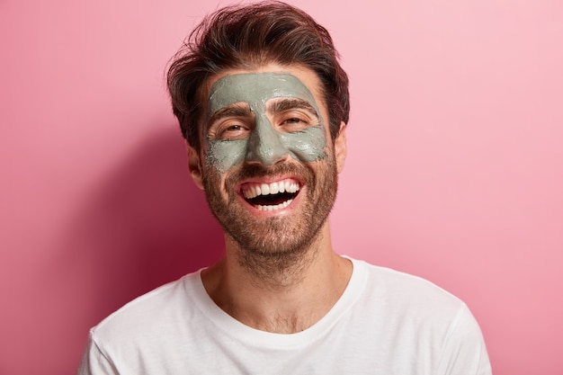 Joyful delighted man has clay mask on face, enjoys spa treatments, has broad smile, being in high spirit, cares about beauty Free Photo