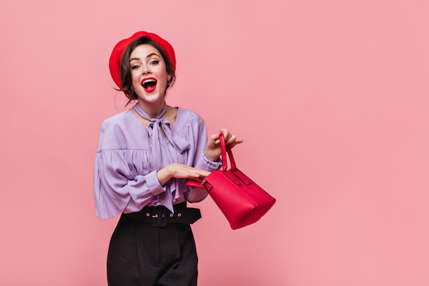 Joyful lady in red hat laughs, holding small bag in her hands on pink background. Free Photo