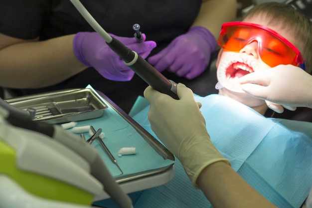 子供の歯科 子供の歯科医の検査赤ちゃんの歯 歯科用椅子での子供の感情 保護用のオレンジ色のメガネとコッファダムの小さな男の子 プレミアム写真