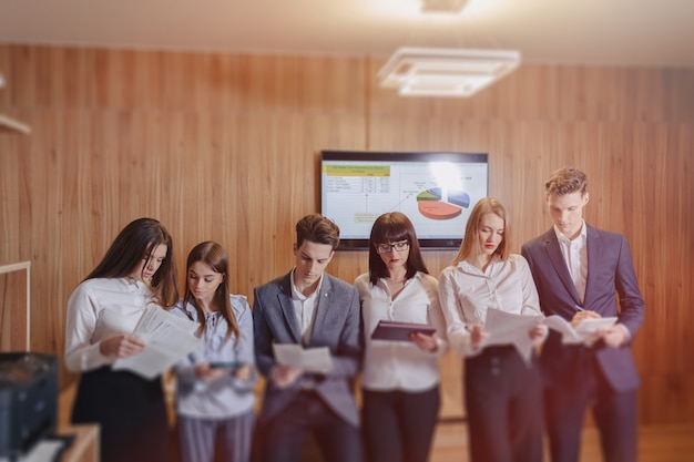 Large team of people is working at one table for laptops, tablets and papers, on the background a large tv set on a wooden wall Premium Photo