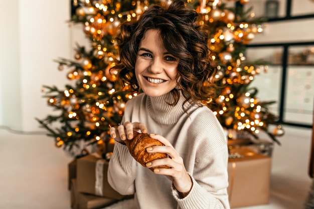 幸せな感情でクリスマスツリーの上にクロワッサンでポーズをとって巻き毛の髪型で幸せな女の子を笑う 新年の朝 クリスマスパーティー 無料の写真