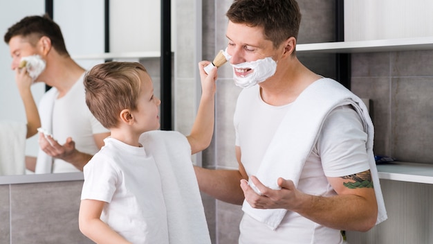 Little boy applying shaving foam on his father's face with brush in the bathroom Free Photo