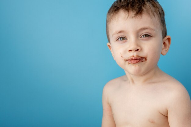 プレミアム写真 青の背景にドーナツチョコレートを食べる少年 かわいい幸せな少年 は彼の口の周りにチョコレートを塗った 子供のコンセプト 子供のためのおいしい食べ物