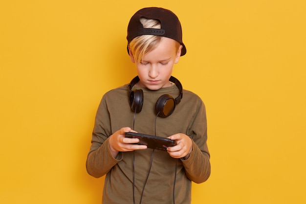 カジュアルな帽子と帽子をかぶって 携帯電話を使用してオンラインビデオゲームをプレイし ヘッドフォンでポーズをとって少し集中している男 無料の写真