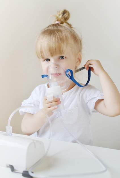 Little girl in a mask for inhalations, making inhalation with nebulizer at home inhaler on the table, indoor, sick child Premium Photo