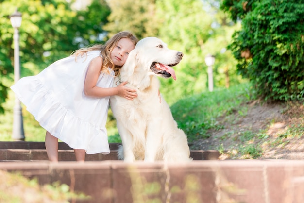 公園で大きな白い犬を持つ少女 白いドレスを着た美しい5歳の女の子が夏の散歩中に彼女のお気に入りの犬を抱擁します プレミアム写真