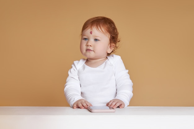 小さな赤い髪の女の赤ちゃんがベージュの背景で遊んでいます 他のみんなのようではなく 額に大きなほくろ プレミアム写真